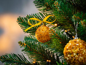 BalticBliss genuine Baltic amber Christmas tree ornament ball with shimmering natural beads and gold ribbon bow hanging on lush green fir branch illuminated by warm fairy lights, handmade in Lithuania, authentic amber Christmas décor ships worldwide