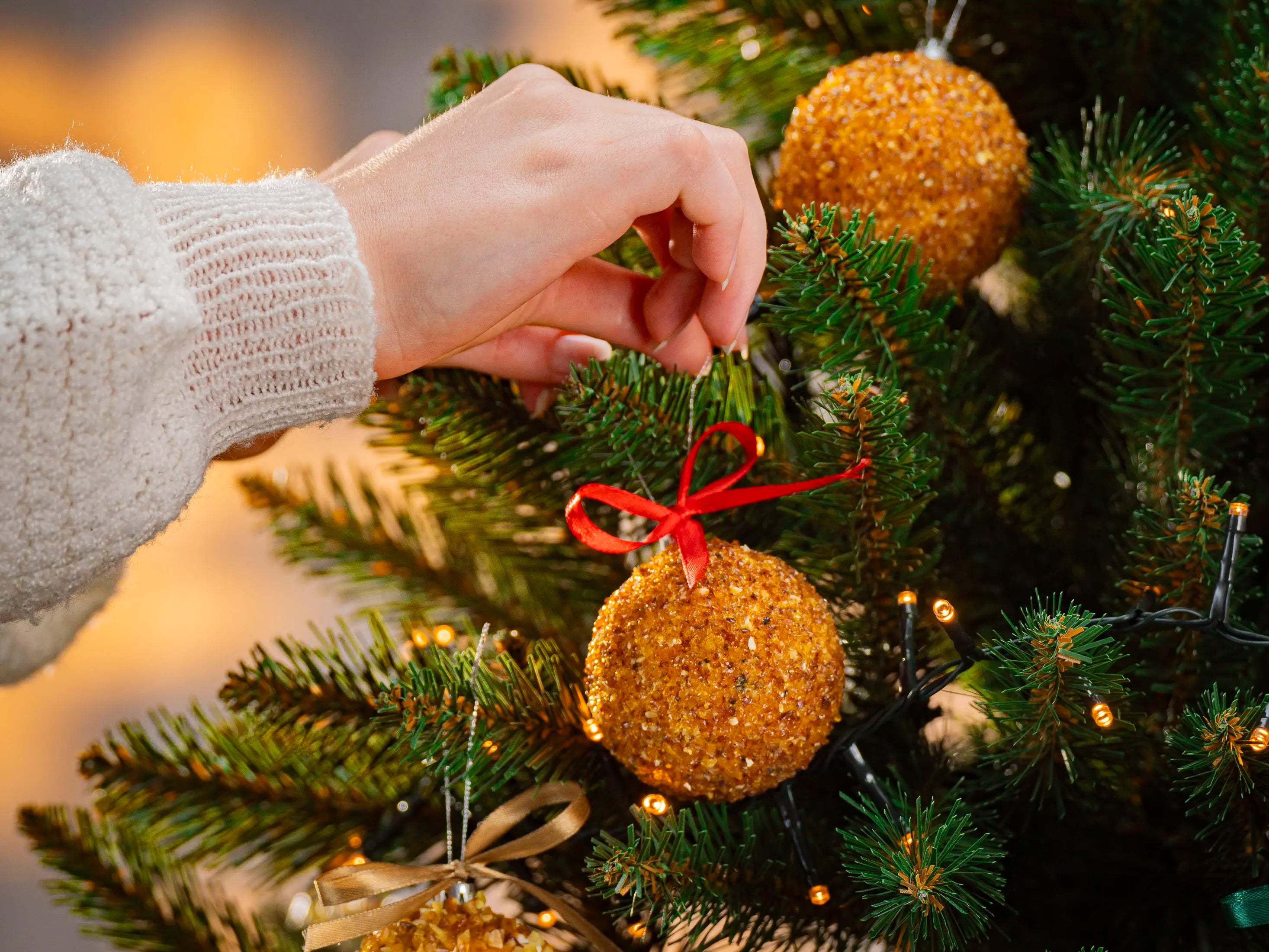 Hand placing BalticBliss genuine Baltic amber Christmas tree ornament ball with shimmering natural beads and red ribbon on lush green fir branch, handmade in Lithuania, authentic amber holiday dΓ©cor ships worldwide