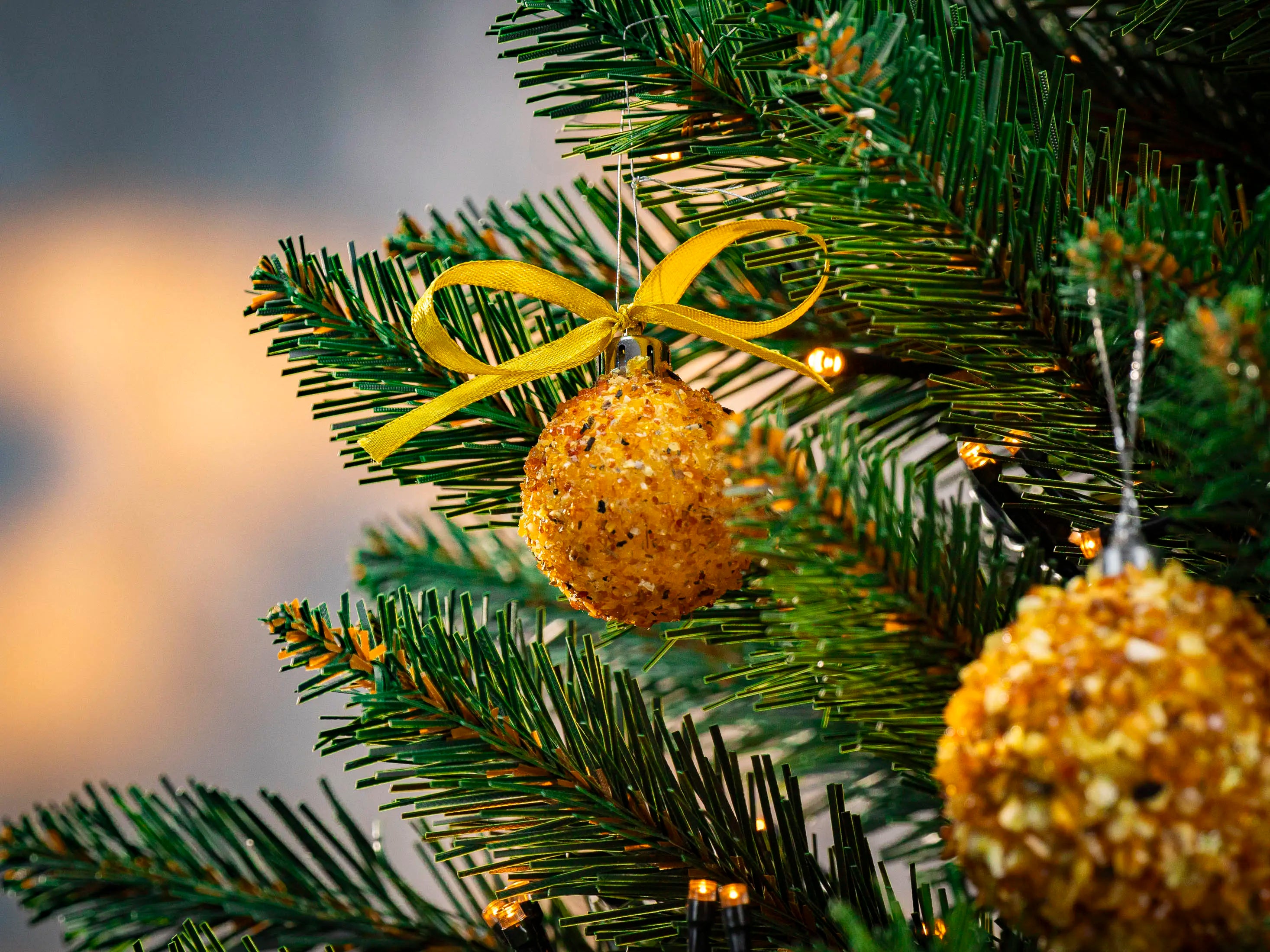 BalticBliss genuine Baltic amber Christmas tree ornament ball with shimmering natural beads and gold ribbon bow hanging on lush green fir branch illuminated by warm fairy lights, handmade in Lithuania, authentic amber Christmas décor ships worldwide